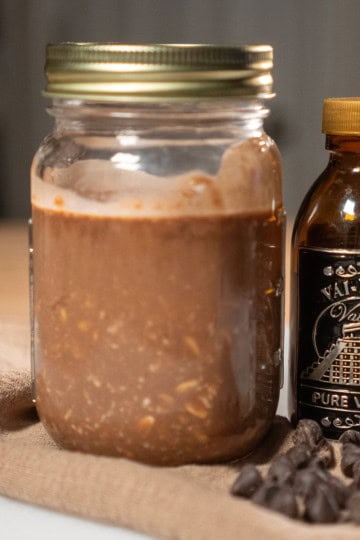 A jar of homemade overnight chocolate oatmeal with vanilla extract, surrounded by chocolate chips on a natural cloth