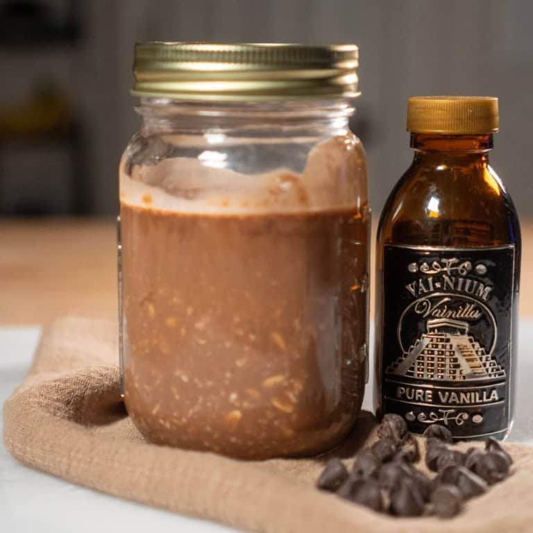 A jar of homemade overnight chocolate oatmeal with vanilla extract, surrounded by chocolate chips on a natural cloth